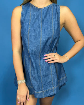 A sleeveless blue denim dress with vertical contrast stitching, modeled against a solid blue background.