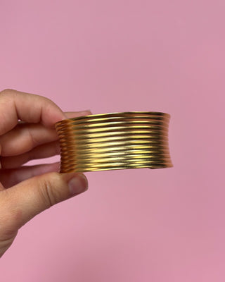 Gold bracelet held by a hand against a pink background
