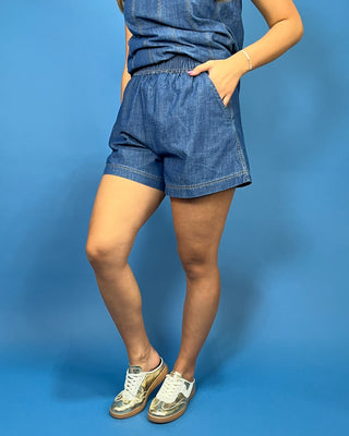 A person modeling blue denim shorts with a matching blue top, standing against a solid blue studio background.