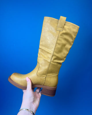 A tan leather knee-high boot with a brown stacked heel, held up against a solid blue background.