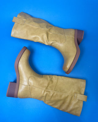A tan leather knee-high boot with a brown stacked heel, held up against a solid blue background.