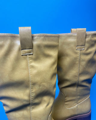 A tan leather knee-high boot with a brown stacked heel, held up against a solid blue background.