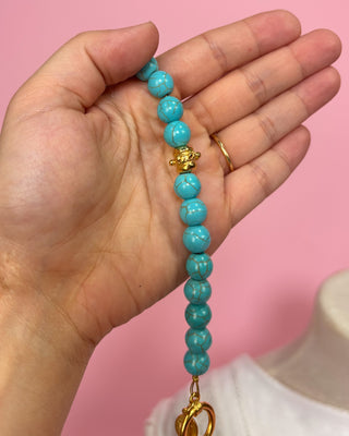 Hand holding a turquoise beaded keychain with a gold clasp against a pink background