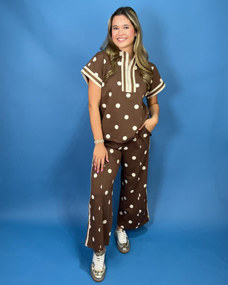 A woman modeling a matching two-piece brown and white polka dot lounge set against a solid blue studio backdrop.