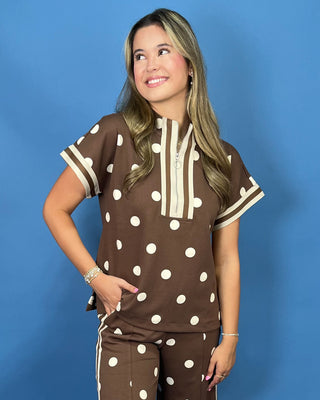 A woman modeling a brown two-piece lounge set with white polka dots and cream stripe accents, standing against a solid blue background.