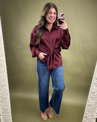 Woman taking a mirror selfie wearing a burgundy shirt and blue jeans on a green background