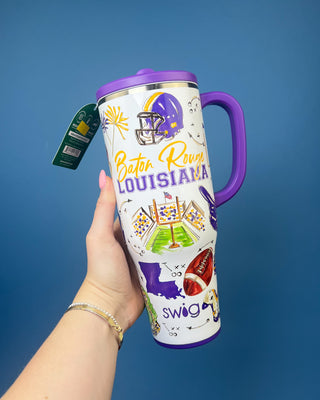 A hand holds a white insulated travel tumbler with a purple lid and handle. The tumbler is decorated with Louisiana Baton Rouge football graphics, including the team name, a helmet, field goals, and the word 'swag'.