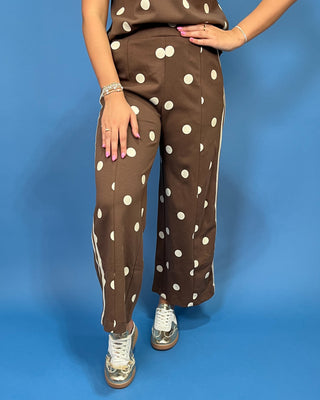 A woman modeling a brown two-piece lounge set with white polka dots, featuring a short-sleeve top with cream trim and matching wide-leg pants