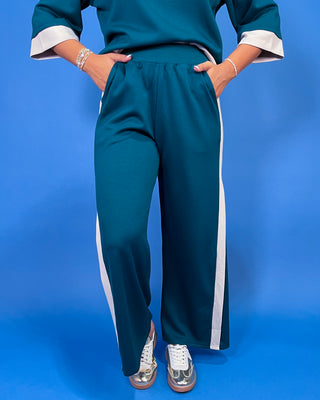 A person wearing teal wide-leg pants with white side stripes and matching teal top with white cuffs, standing against a blue studio background.