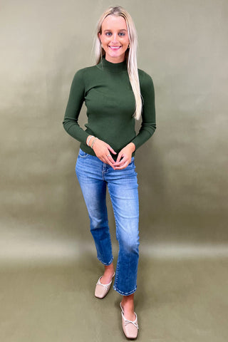 Woman wearing a green long-sleeve top and blue jeans against a plain background