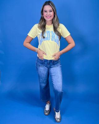 A woman posing in a light yellow graphic t-shirt with blue lettering, paired with blue jeans and silver sneakers, set against a solid blue studio background.