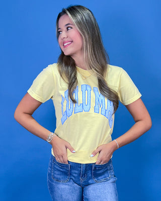 A woman posing in a light yellow graphic t-shirt with blue lettering, paired with blue jeans
