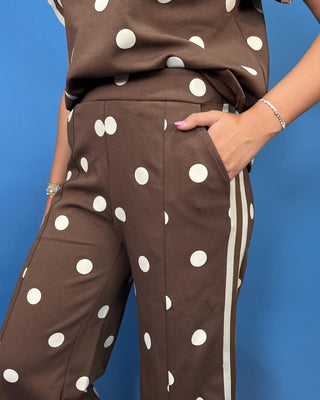 A close-up of a person wearing brown wide-leg pants with large white polka dots and white side stripes, paired with a matching brown top,