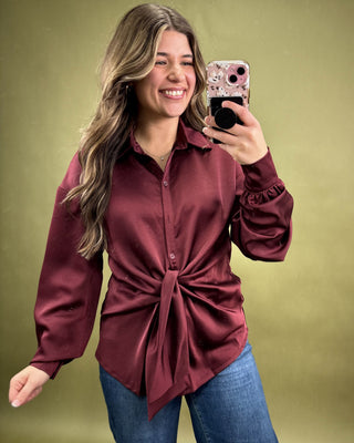 Woman taking a selfie wearing a burgundy shirt with a tie detail and blue jeans on a green background.
