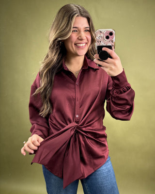 Woman taking a selfie wearing a burgundy knotted blouse against a green background