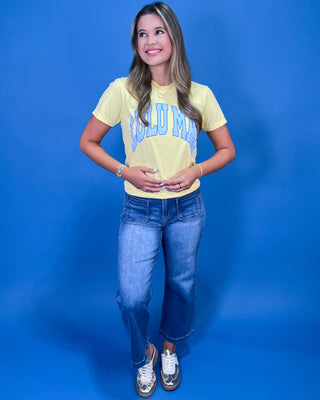 A woman posing in a light yellow graphic t-shirt with blue lettering, paired with blue jeans