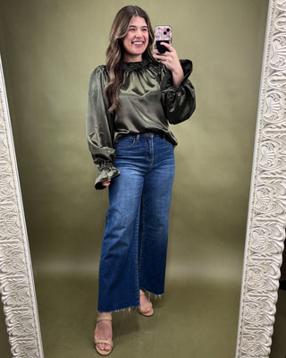 Woman taking a mirror selfie wearing a green blouse and blue jeans on a green background.
