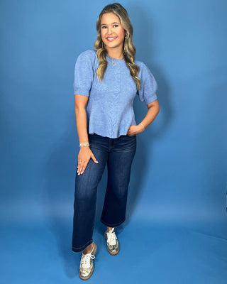 Woman modeling a light blue cable knit puff sleeve peplum sweater top with dark wash wide leg jeans.