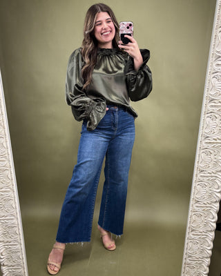 Woman taking a mirror selfie wearing an olive green velvet top and blue jeans on a green background.