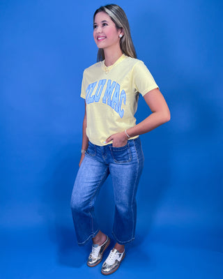 A woman posing in a light yellow graphic t-shirt with blue lettering, paired with blue jeans