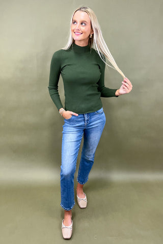Woman wearing a green turtleneck and blue jeans against a plain background
