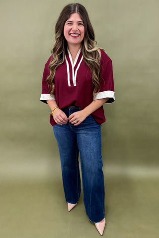 Woman wearing a burgundy tunic top with white trim and wide sleeves, paired with dark wash wide leg jeans and nude pointed heels, boutique fashion photography on olive backdrop