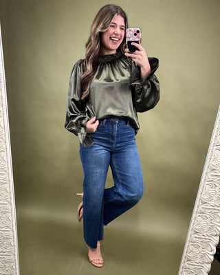 Woman taking a mirror selfie wearing a green velvet top and blue jeans on a green background.