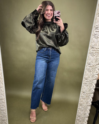 Woman taking a mirror selfie wearing an olive green velvet top and blue jeans on a green background.