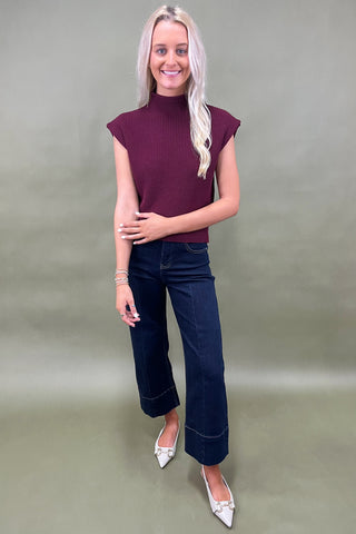 Woman wearing a maroon sleeveless sweater and blue jeans against a plain background