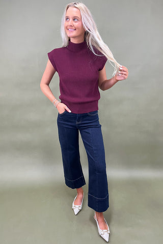 Woman wearing a purple sleeveless sweater and blue jeans against a plain background