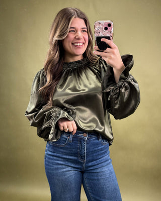 Woman taking a selfie wearing a green satin blouse and blue jeans on a gold background