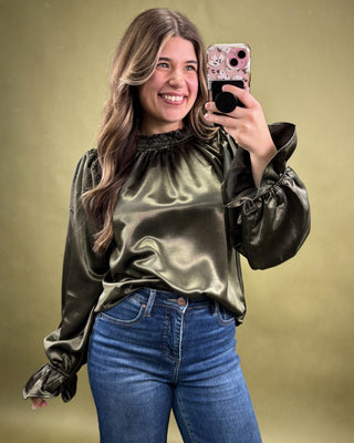 Woman taking a selfie wearing a metallic green blouse and blue jeans on a gold background