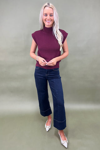 Woman wearing a maroon sleeveless sweater and blue jeans against a plain background