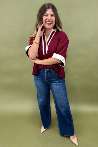 Woman wearing a burgundy tunic top with white trim and wide sleeves, paired with dark wash wide leg jeans and nude pointed heels, boutique fashion photography on olive backdrop