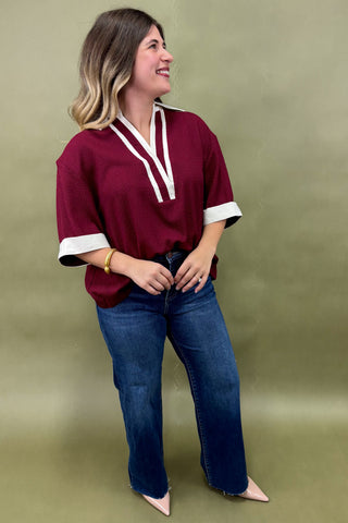 Woman wearing a burgundy tunic top with white trim and wide sleeves, paired with dark wash wide leg jeans and nude pointed heels, boutique fashion photography on olive backdrop