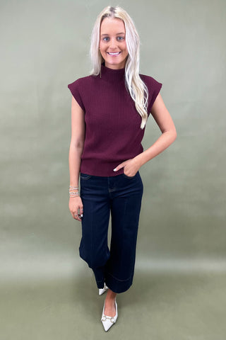 Woman wearing a maroon sleeveless sweater and blue jeans against a plain background