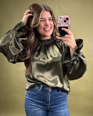 Woman taking a selfie wearing a metallic green blouse and blue jeans on a gold background