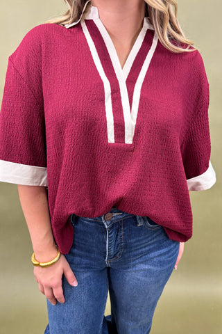 Woman wearing a burgundy tunic top with white trim and wide sleeves, paired with dark wash wide leg jeans and nude pointed heels, boutique fashion photography on olive backdrop