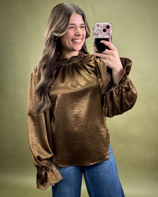 Woman wearing a bronze long-sleeve top and blue jeans, taking a selfie against a green background.