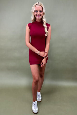 Woman wearing a sleeveless burgundy bodycon mini dress styled with white sneakers, boutique fashion photography on olive backdro