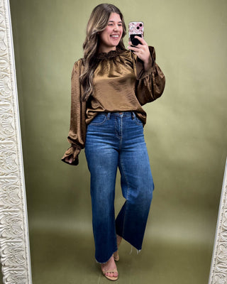 Woman taking a mirror selfie wearing a brown blouse and blue jeans on a green background