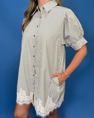 A woman wearing a short-sleeve button-front shirt dress with thin vertical black stripes on a cream background and white lace trim at the hem and cuffs