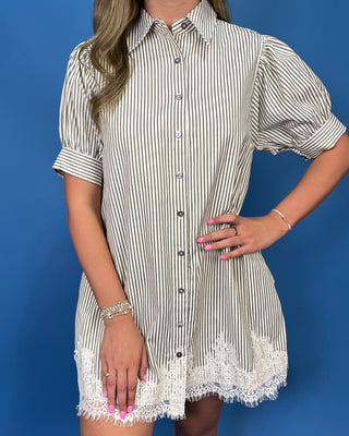 A woman wearing a short-sleeve button-front shirt dress with thin vertical black stripes on an off-white base and white lace trim at the hem
