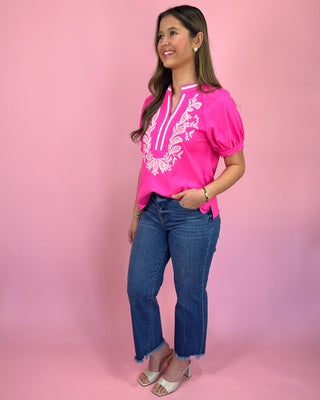 Woman wearing a bright pink top with white embroidery and blue jeans on a pink background