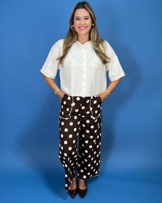 A woman modeling a white short-sleeve button-up blouse and brown wide-leg pants with white polka dots, standing against a solid blue studio backdrop.