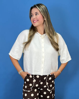 A woman wearing a white short-sleeve button-up blouse and brown polka-dot pants, standing against a solid blue background.