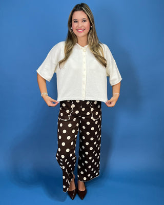 A woman modeling a short-sleeve cream button-up blouse paired with brown wide-leg pants featuring a white polka-dot pattern, standing against a solid blue studio backdrop.