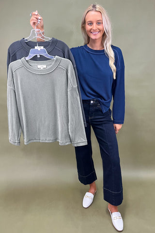 Woman holding a gray ribbed sweater next to a blue sweater against a plain background