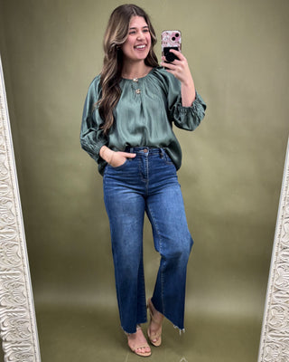 Woman taking a mirror selfie wearing a green blouse and blue jeans on a green background