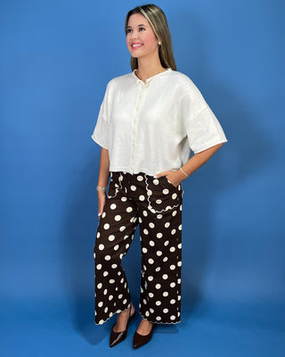A woman modeling a white short-sleeve button-front blouse and brown wide-leg pants with white polka dots, standing against a solid blue studio background.